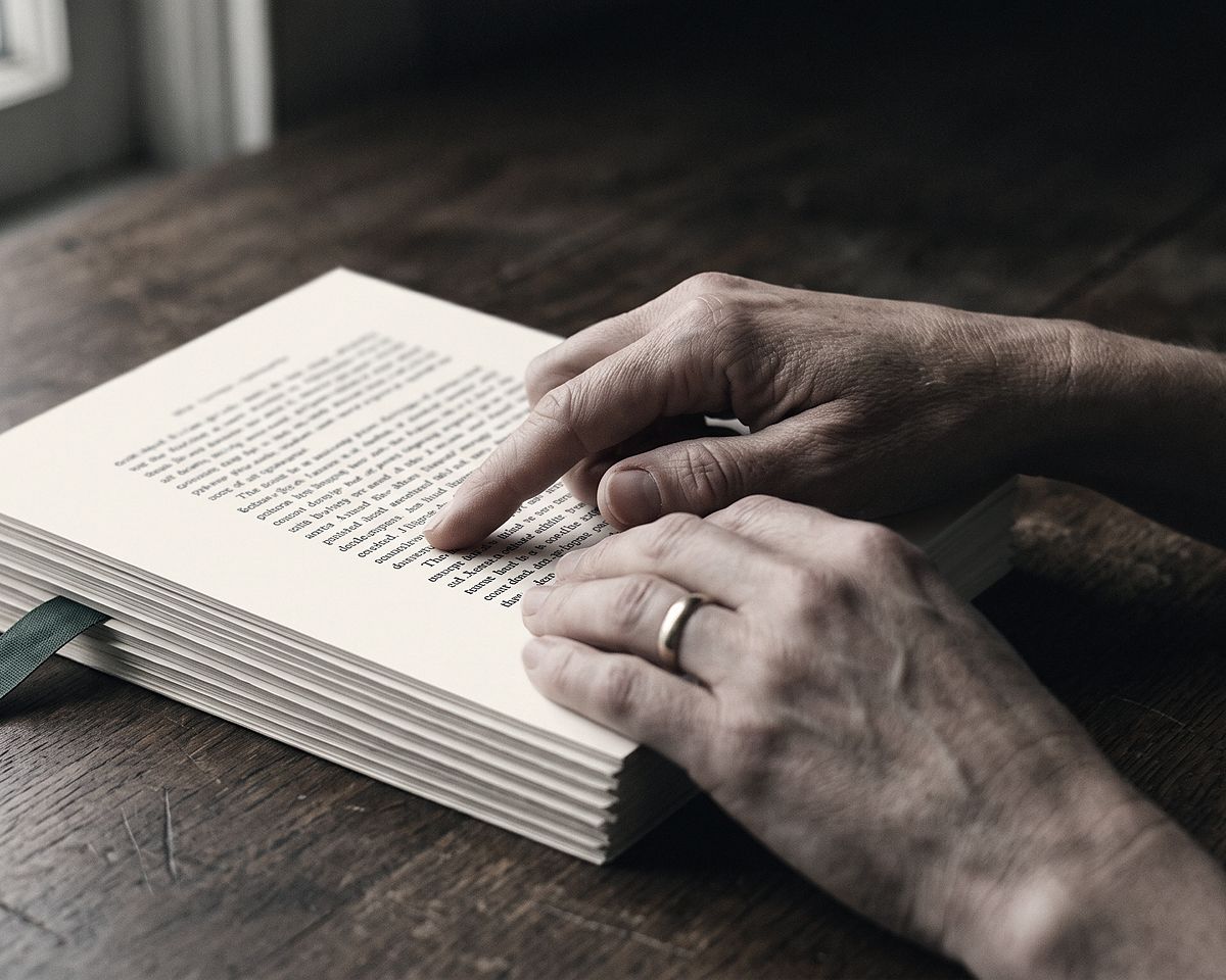 Hands resting on a stack of printed pages on a wooden desk, one finger paused partway down a single line.
