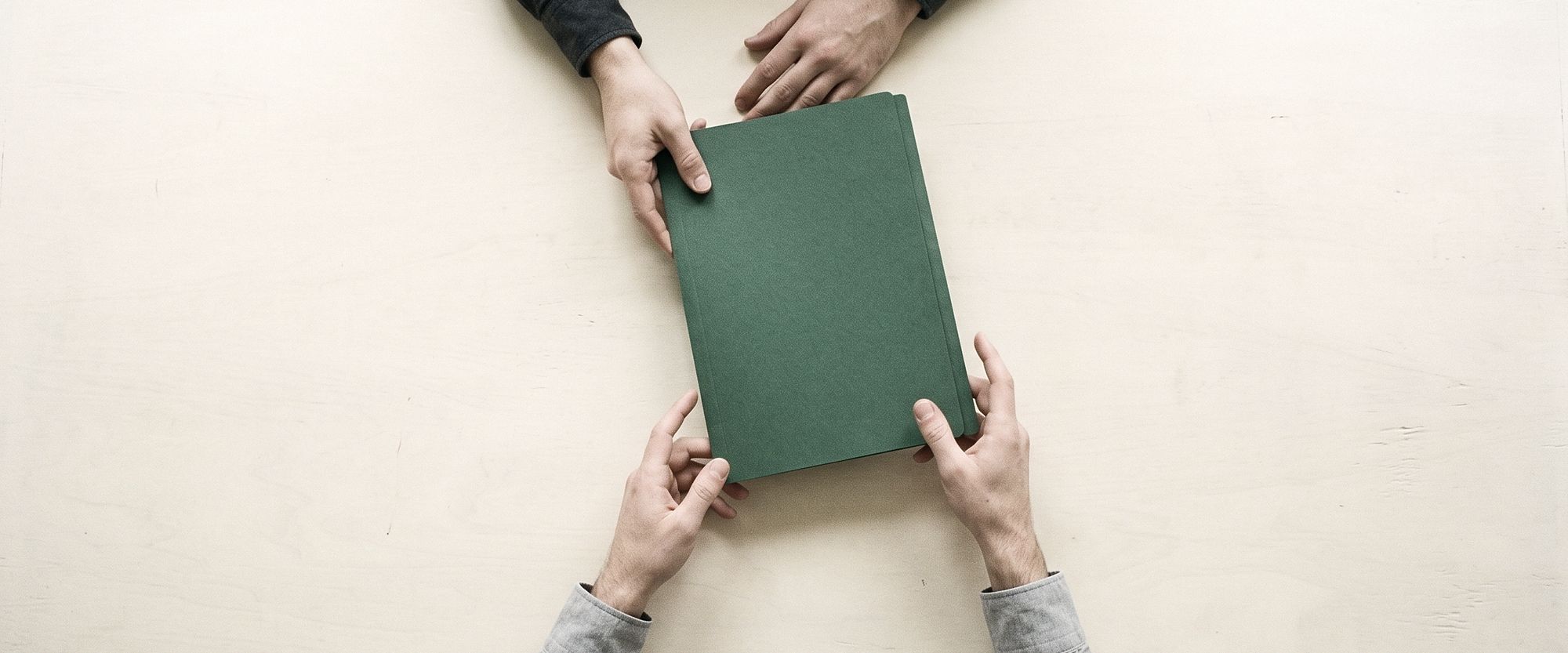 Seen from above, one person slides a plain closed folder of documents across a pale table toward another.